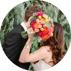 photo of a couple leaning in to kiss while holding a brightly-colored bouquet in front of their faces